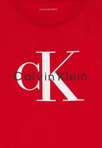 Czerwona koszula z dużymi białymi inicjałami "cK" i mniejszym czarnym napisem "Calvin Klein" nadrukowanym z przodu, widocznym okrągłym dekoltem.