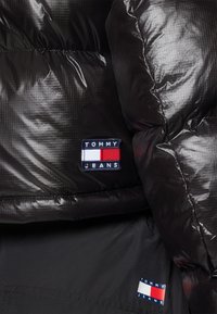 Chaqueta de plumas negra con una textura brillante y acolchada. Incluye un parche rectangular con el logo "TOMMY JEANS" en azul marino, rojo y blanco.