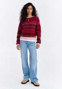 Czerwony i różowy sweter w paski z dzianiny z prążkowanym wykończeniem, zestawiony z jasnoniebieskimi dżinsami o szerokich nogawkach i beżowymi butami. Model stoi na białym tle.