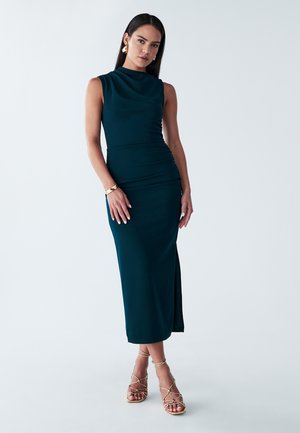Robe midi sans manches teal, avec une silhouette ajustée, une fente sur le côté et une texture froncée. Modèle portant des sandales dorées à lanières et des bijoux.
