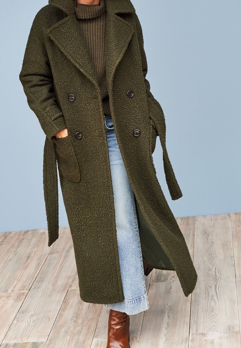 Personne portant un long manteau texturé vert foncé par-dessus un pull marron, un jean bleu clair et des bottes en cuir marron sur un sol en bois.