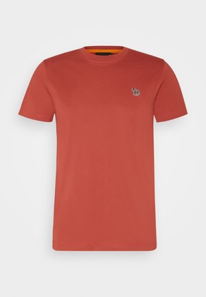 Person iført en rustfarvet t-shirt med et lille zebra-logo og sorte bukser, stående ved siden af en brun læderstol mod en lys væg.