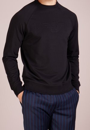 Mężczyzna w czarnej bluzie z okrągłym dekoltem z wytłoczonym logo oraz granatowych spodniach z cienkimi, pionowymi niebieskimi paskami, z rękami w kieszeniach.