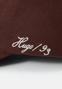 Brown cap featuring textured fabric with stitched white "Hugo / 93" lettering on the side, showcasing a classic curved shape.