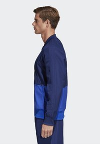 Blaues Sportjackett mit einem zweifarbigen Design, bestehend aus einem dunkleren oberen Abschnitt und einem helleren unteren Teil. Aus glattem Stoff gefertigt, mit Reißverschlusstaschen.