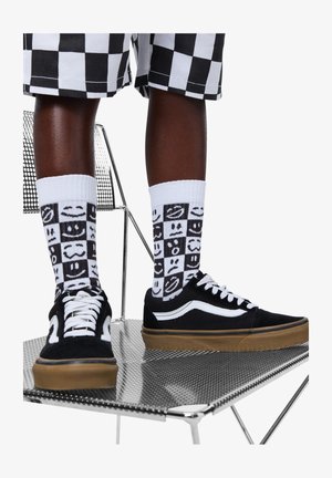 Μαύρα καμβά παπούτσια με άσπρα στοιχεία, συνδυασμένα με άσπρες κάλτσες με μοτίβο χαμογελαστών προσώπων και καρό σορτς. Φάση με μεταλλική καρέκλα στο φόντο.