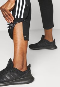 Svarta sportbyxor med vita ränder, tryckknapps stängning vid nederkanten och en strömlinjeformad passform. Bärs med svarta sportskor.