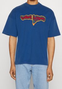 Blaues Baumwoll-T-Shirt mit einem mehrfarbigen, gestickten Logo auf der Brust. Kurze Ärmel und lässige Passform. Minimalistisches Design.