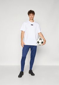 Camiseta branca com pequeno logotipo no peito, combinada com calças desportivas azuis-marinho com riscas brancas nas laterais. Segura uma bola de futebol preta e branca.