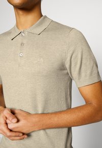 Beige pikétröja i mjukt stickat material, med klassisk krage, treknappsgylf och liten broderad logotyp på bröstet.