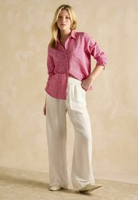 Ružová textúrovaná košeľa s gombíkmi a jedným vreckom na hrudi, v kombinácii s voľne strihanými bielymi nohavicami a svetlými topánkami.