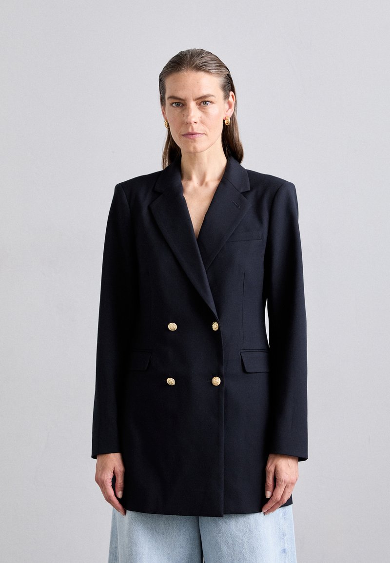 Marineblå, dobbeltradet blazer med gullknapper, utskårne lapeller og sidelommer; glatt stoff med strukturert form og litt oversized passform.