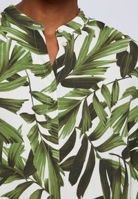 Chemise à manches courtes avec un col, fabriquée en tissu léger, ornée d'un motif de feuilles tropicales vertes sur fond blanc.