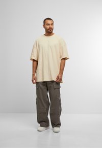 Cămașă bej oversized cu mânecă scurtă din material texturat, combinată cu pantaloni cargo largi, gri închis, cu multiple buzunare și o croială lejeră.
