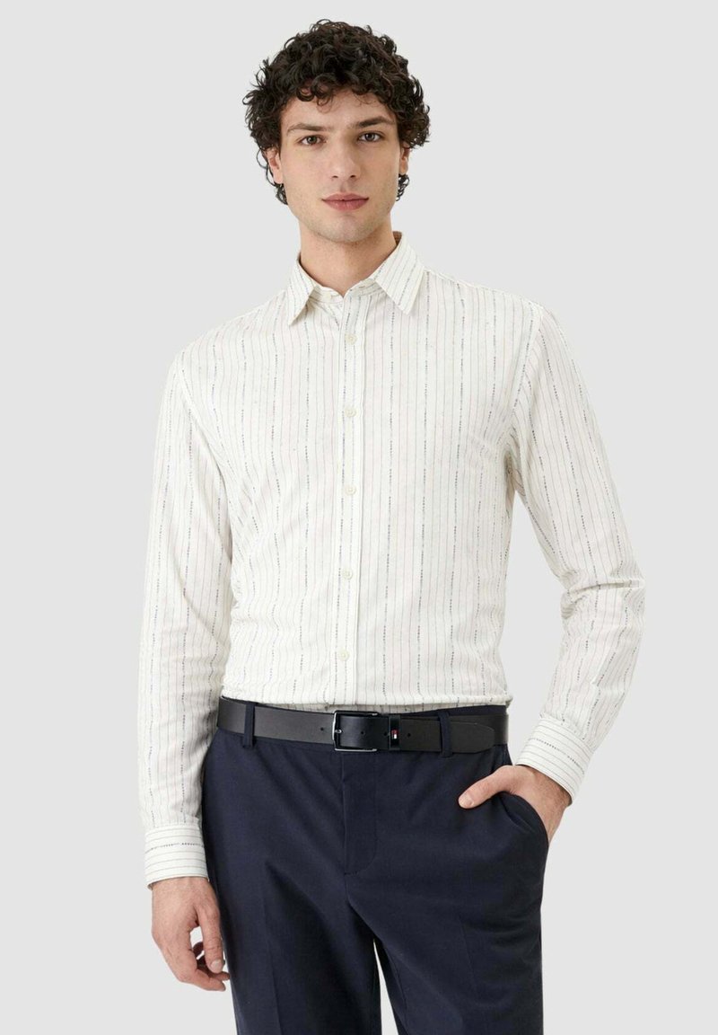 Homme aux cheveux foncés bouclés portant une chemise blanche à rayures boutonnée, rentrée dans un pantalon bleu marine avec une ceinture noire, debout avec une main dans la poche.