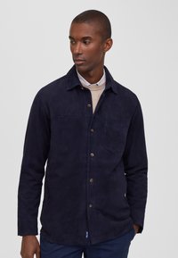 Camisa de ante azul marino con cierre de botones en la parte delantera, cuello de punta, bolsillo en el pecho y mangas largas. Presenta una pequeña etiqueta de la marca en el dobladillo.