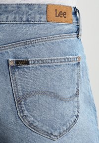Ljusblå denimjeans med en läder Lee-logotyp på midjan, kontrasterande sömmar och en sydd bakficka med en böjd design.