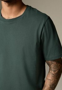 Tricou din bumbac verde, cu guler rotund și mâneci scurte, având o textură netedă și detalii de cusătură subtile.