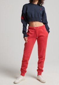 Person wearing a cropped navy sweatshirt and red drawstring sweatpants with white sneakers, standing with one hand in pocket.