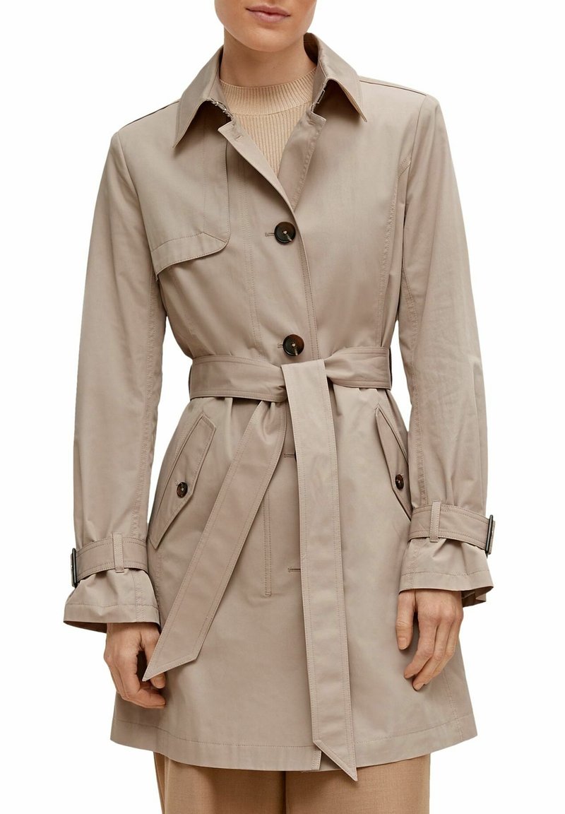 Femme portant un trench-coat beige ceinturé avec des boutons foncés, des poches à rabat et des sangles ajustables aux manches, par-dessus un pull marron clair.