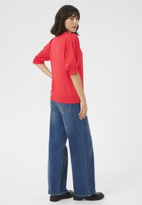 Pull rouge à manches bouffantes 3/4, associé à un jean bleu taille haute à jambes larges. Texture lisse, ourlet côtelé et accents minimalistes.