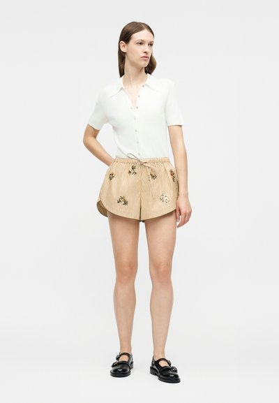 Γυναίκα με λευκό κοντομάνικο πουκάμισο με κουμπιά, μπεζ ριγέ σορτς με κέντημα λουλουδιών και μαύρα παπούτσια με αγκράφα, στέκεται με το ένα χέρι στην τσέπη.