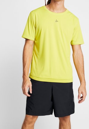 Mężczyzna ubrany w limonkową sportową koszulkę z krótkim rękawem i czarne sportowe spodenki na jednolitym szarym tle.