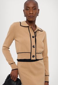 Beige knitted sweater with a collar, black trim, and six buttons. Texture appears soft; features a fitted design and contrasting lines.