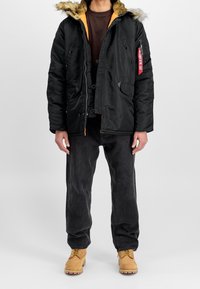 Chaqueta negra acolchada con capucha forrada de piel, forro naranja, cierre de botones y cremallera, múltiples bolsillos y un parche en la manga.