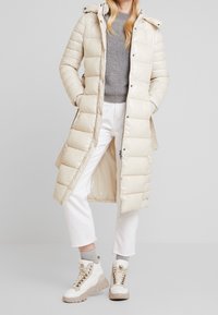 Abrigo largo beige acolchado con exterior brillante, bolsillos con cremallera y capucha. Lleva puesto un suéter gris y pantalones cortos blancos, combinados con botas beige.