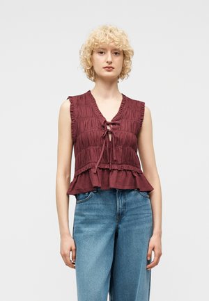 Ung kvinne med kort krøllete blondt hår iført ermeløs mørkerød bluse med volanger og blå jeans med høyt liv, stående foran en ensfarget bakgrunn.