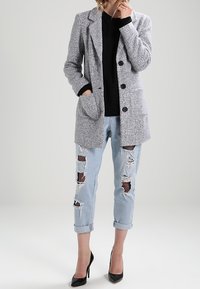 Šedý blazer z vlněné směsi s dvěma předními kapsami a třemi černými knoflíky, spárovaný se světlými modrými džíny s opotřebením, které mají akcenty z rybářské sítě.