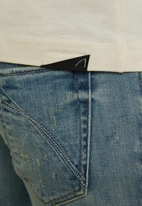 Kermat t-paita, jossa on musta tunniste helman kohdalla, yhdistettynä kulutettuihin vaaleansinihin farkkuihin, joissa on tikkausyksityiskohtia ja haalistunut pinta.