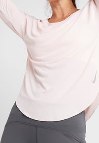 Lichtroze sporttop van een gladde stof, met een ronde halslijn en een gebogen zoom. Bevat een subtiele grijze Nike-logo aan de zijkant.
