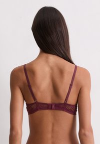 Donna vista di spalle che indossa un reggiseno in pizzo bordeaux con spalline regolabili su uno sfondo chiaro e semplice.