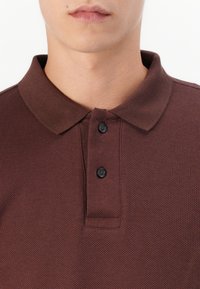 Cămașă polo maro cu țesătură texturată, guler clasic și două butoane negre pe plachetă. Vedere detaliată a zonei gâtului.