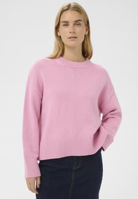 Femme blonde portant un pull en tricot rose à manches longues et un jean bleu foncé, une main dans la poche, sur un fond uni.