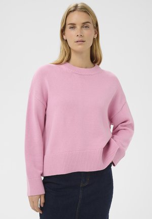Blonde Frau trägt einen langärmligen pinkfarbenen Strickpullover und dunkelblaue Jeans, eine Hand in der Tasche, vor einem einfarbigen Hintergrund.