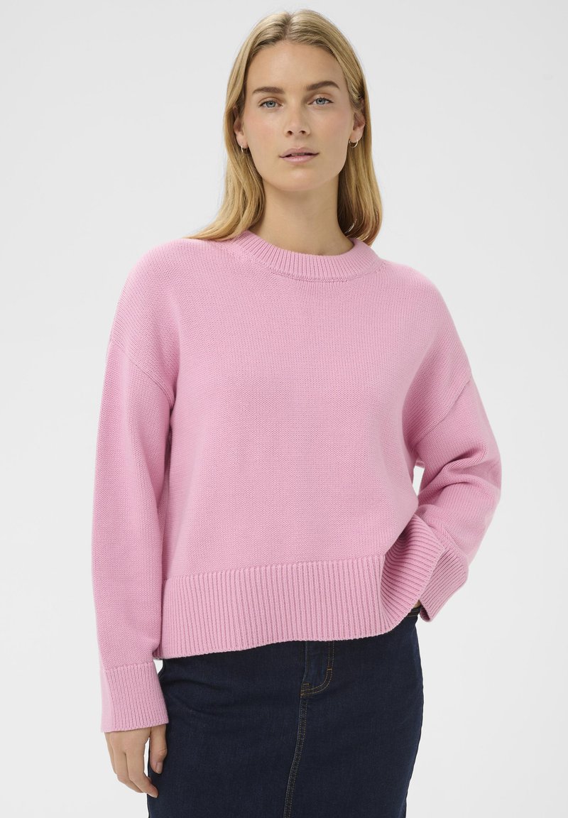 Femme blonde portant un pull en tricot rose à manches longues et un jean bleu foncé, une main dans la poche, sur un fond uni.