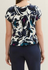 Maglietta a maniche corte con un motivo floreale in blu navy, verde teal e magenta su sfondo bianco, realizzata in un tessuto morbido e con una vestibilità ampia.