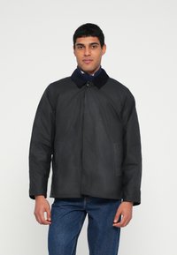 Casaco preto com gola em azul escuro, com fecho de botões na frente e dois bolsos laterais. O tecido aparenta ser suave com textura mínima.