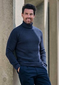 Mann mit dunklen Haaren und Bart, der an einer Betonwand lehnt, einen marineblauen Rollkragenpullover und passende Hose trägt, die Hände in den Taschen, lächelnd.