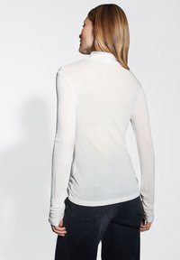 Femme aux cheveux châtains clairs portant un haut à col roulé blanc à manches longues et un pantalon foncé, de dos contre un fond uni.