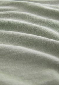 Svetlo zelena tkanina z mehko, gladko teksturo ter valovitimi gubami, ki razkrivajo fino pleten vzorec in subtilen lesk. Poudarja mehkobo in padanje tkanine.