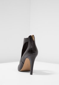 Musta korkokenkä, jossa on sileä nahkapinta, takazippari, sivuilla leikkaukset ja siro korko. Alaosassa näkyy beige pohja.