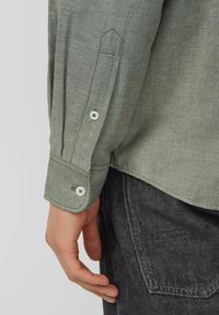 Detail van een groen shirt met lange mouwen en een geruite manchetknoop, met witte knopen en een getextureerde stof, met een zichtbare zak op zwarte broek.