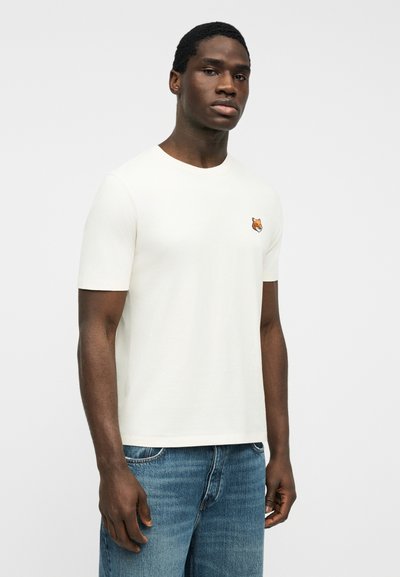 Νέος άνδρας που φοράει λευκό κοντομάνικο T-shirt με μικρό λογότυπο κεφαλής αλεπούς στο στήθος και μπλε τζιν, στέκεται μπροστά σε απλό φόντο.