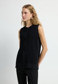 Schwarzes ärmelloses Oberteil mit strukturiertem Rippenmuster, Knopfleiste, seitlichen Schlitzen und geradem Schnitt, kombiniert mit schwarzen Hosen.