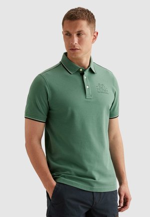 Mężczyzna w zielonej koszulce polo z krótkim rękawem i zapiętym kołnierzykiem oraz ciemnych szortach, stoi z jedną ręką w kieszeni, patrząc w lewo.