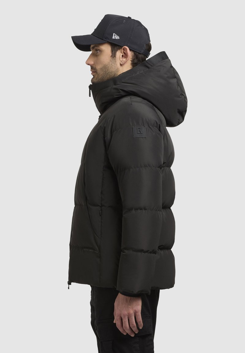 Mann in schwarzer Steppjacke mit Kapuze und schwarzem Baseballcap, der im Profil vor einem schlichten hellen Hintergrund steht.